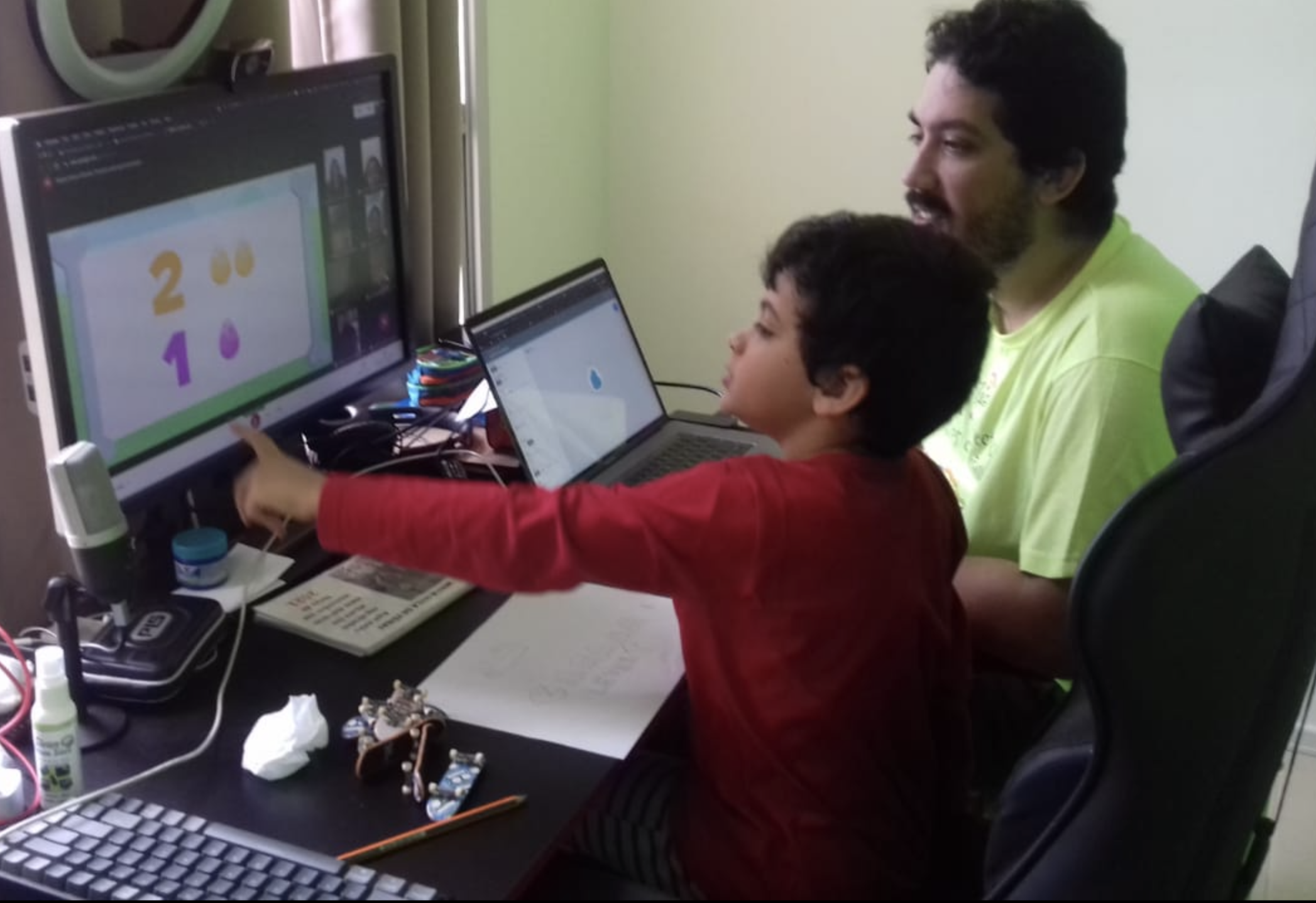 Pai e Miguel lado a lado em frente ao computador, testando um jogo educacional durante uma aula virtual no período da pandemia